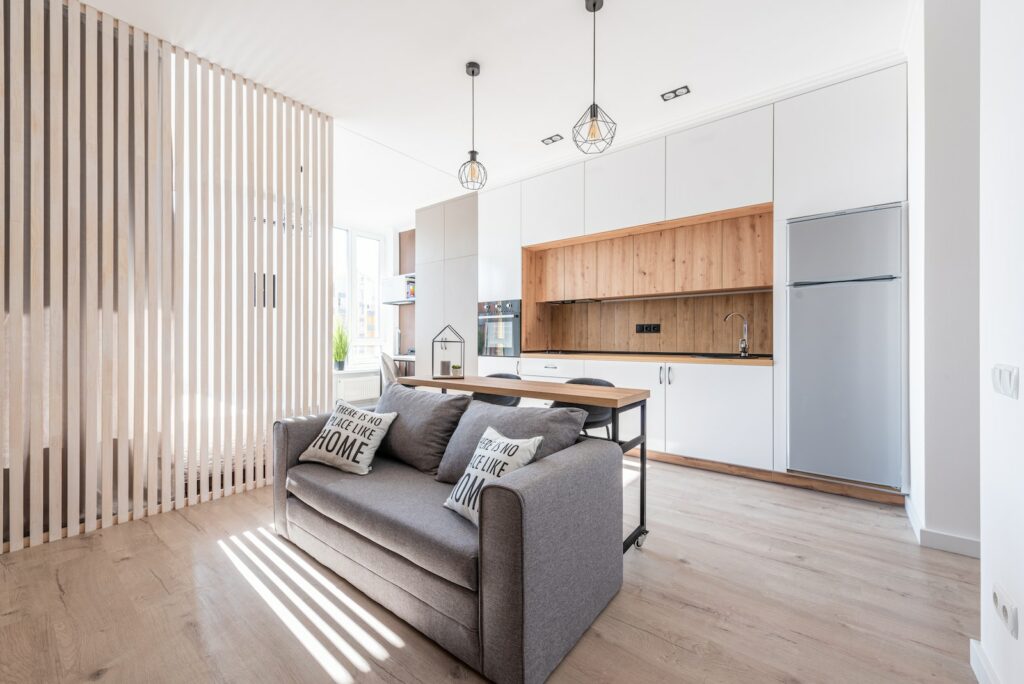 Interior of contemporary open plan kitchen with white cabinets and cozy sofa with cushions in sunlight